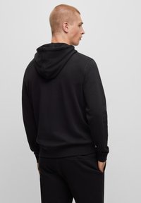 Giovane uomo con capelli corti, che indossa un hoodie nero e pantaloni neri abbinati, in piedi di lato su uno sfondo chiaro e semplice.