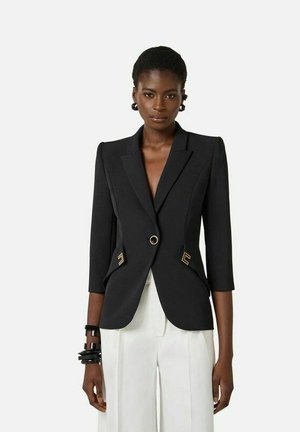 Donna che indossa un blazer nero aderente con dettagli dorati e pantaloni bianchi a vita alta, in piedi davanti a uno sfondo semplice.