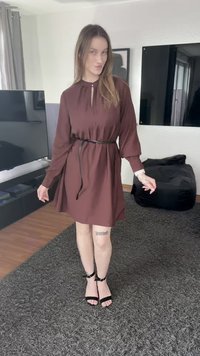 Vestido castanho de manga comprida com decote em gota, cintura marcada por um cinto preto. Usado com sandálias pretas com tiras, sobre um tapete texturizado.