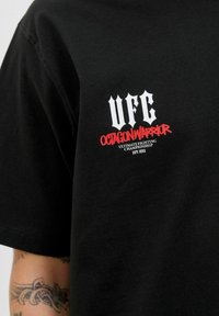 Camisa negra con el texto blanco y rojo "UFC Octagon Warrior Ultimate Fighting Championship EST. 1993" en el pecho, brazo tatuado parcialmente visible.