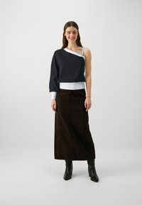 Sweatshirt asymétrique noir avec une manche longue et des finitions blanches, associé à une jupe en denim marron foncé. Le modèle porte des bottines noires.