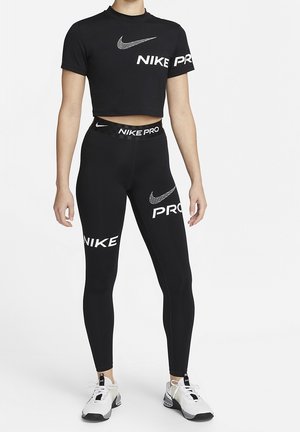 Personne portant un t-shirt court noir Nike Pro et des leggings avec des logos Nike blancs ainsi que des chaussures de sport blanches, debout sur un fond blanc.