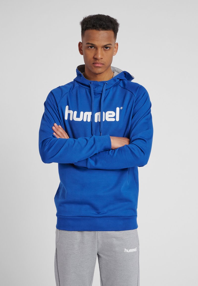 Junger Mann steht mit verschränkten Armen, trägt einen blauen Hummel-Hoodie und graue Hummel-Jogginghosen vor einem einfarbigen Hintergrund.
