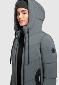 Giacca a vento grigia con cappuccio, chiusura con zip e cordini elastici. Presenta una texture trapuntata e una patch con logo sulla manica.