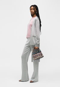Femme aux longues tresses portant un haut rose, un cardigan gris et un pantalon, tenant un sac à main à motifs zigzag multicolores, debout de profil.