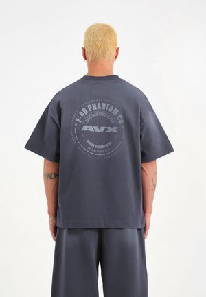 Persona con cabello rubio corto, vistiendo una camiseta de gran tamaño gris oscuro y pantalones cortos, de espaldas a la cámara, la camiseta tiene un gráfico circular con texto.