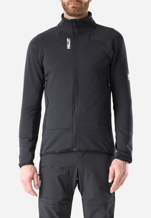 Man wearing black zip-up fleece jacket and black pants with zippered pocket, standing against a white background.