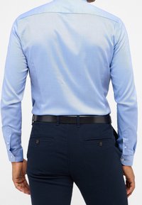 Chemise bleu clair à manches longues, dotée d'un léger éclat, associée à un pantalon texturé bleu marine foncé et à une ceinture noire.
