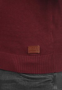 Burgunderfarbener Strickpullover mit geripptem Saum, aus strukturiertem Stoff und mit einem Lederschild, auf dem "BLEND" eingeprägt ist.