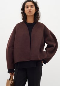 Rudas per didelis švarkas, pagamintas iš minkštos medžiagos, su V formos iškirpte ir nuleistais pečiais, derinamas su juodu raštuotu megztiniu ir juodomis kelnėmis.