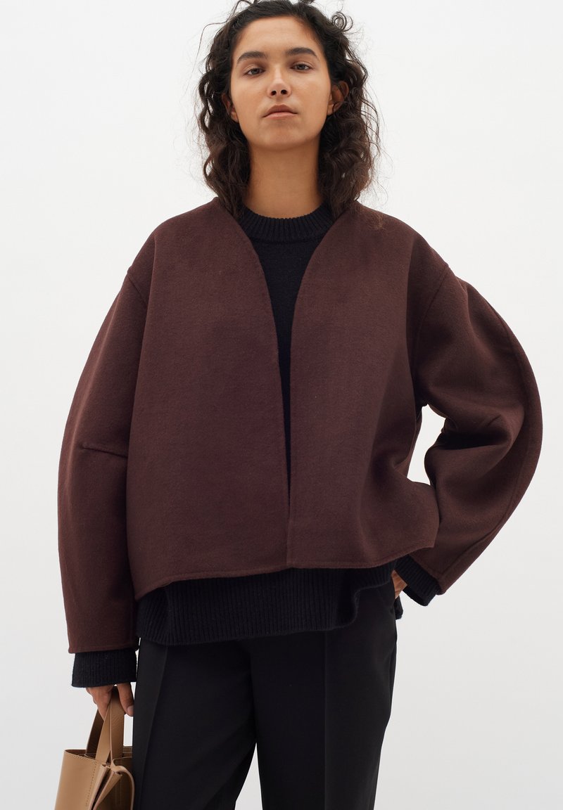 Rudas per didelis švarkas, pagamintas iš minkštos medžiagos, su V formos iškirpte ir nuleistais pečiais, derinamas su juodu raštuotu megztiniu ir juodomis kelnėmis.