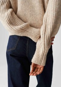 Persona che indossa un maglione a coste color crema con maniche lunghe e jeans di denim blu scuro, con le mani incrociate dietro la schiena.