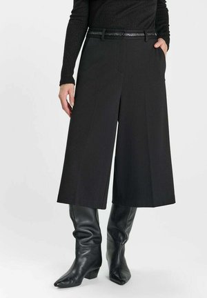 Vrouw die zwarte wijde cropped broek met riem draagt, een zwarte top met lange mouwen en zwarte leren kniehoge laarzen, hand in zak.
