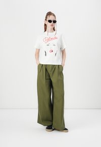 White short-sleeve t-shirt featuring a graphic design with red text "Blondie." Dark green wide-leg pants, paired with black shoes. Black sunglasses.
