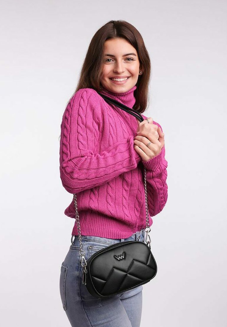 Černá prošívaná crossbody taška s řetízkovým popruhem, se zakřiveným tvarem a embosovaným logem. Nosená s růžovým sveterem z pleteniny.
