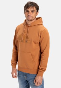 Brun hættetrøje lavet af blødt materiale, med en frontlomme og et logo grafisk design. Parret med blå denim jeans.