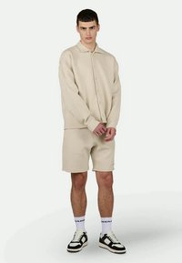 Beige gebreide short-sleeve shirt met knopen, gecombineerd met bijpassende shorts. Details omvatten een textuurstof en zwart-witte sneakers.