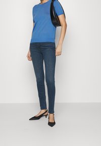 Mujer vistiendo un top azul de manga corta, jeans ajustados oscuros, tacones de tipo slingback negros y un bolso negro de hombro, frente a un fondo liso.