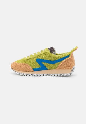 Basket basse avec tige en suède texturé vert et beige, détail zigzag bleu, lacets blancs et semelle blanche tachetée.