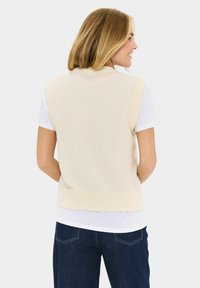 Blonde vrouw die glimlacht, gekleed in een crèmekleurige mouwloze sweatervest over een wit T-shirt en donkerblauwe jeans, met de rug naar de kijker en handen achter haar rug.