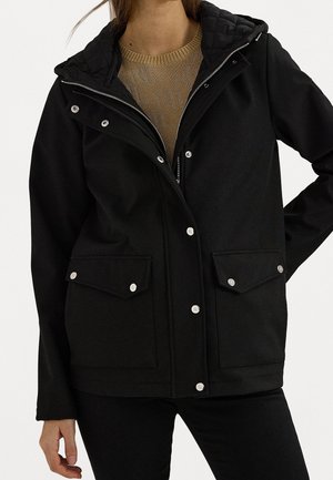 Žena v černé bundě s kapucí, předním zipem a kapsami na druky přes béžový pletený svetr.