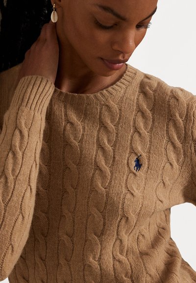 Pull en tricot beige avec un col rond et des manches longues, doté d'un petit patch logo bleu sur la poitrine gauche. Texture visible.