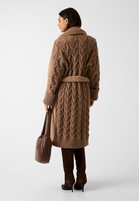 Brązowy kardigan dzianinowy z haftowanym wzorem, ściągaczami przy mankietach i pasującym paskiem, noszony na brązowych kozakach do ud oraz z brązową torbą.