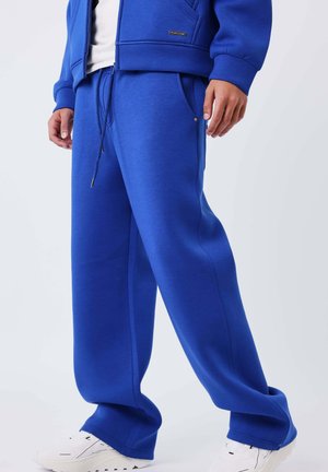 Hombre vistiendo pantalones deportivos azul brillante con cintura ajustable y zapatillas blancas, de pie contra un fondo claro y sencillo.