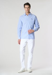 Camicia azzurra chiara a maniche lunghe con un piccolo logo sul petto, abbinata a pantaloni bianchi e sneaker bianche su uno sfondo neutro.