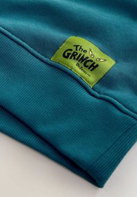 Poignet en tissu bleu sarcelle avec un patch vert cousu montrant le texte "The Grinch" et deux yeux étroits et malicieux au-dessus du texte.