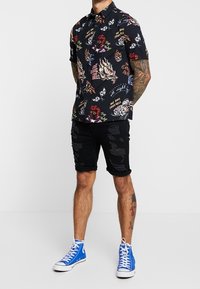 Camisetta nera a maniche corte con stampa grafica colorata, abbinata a pantaloni corti neri strappati e scarpe blu alte.