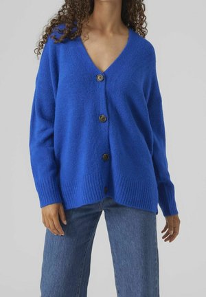 Femme portant un cardigan boutonné bleu vif et ample avec des poignets et un ourlet côtelés, associé à un jean bleu de lavage moyen.