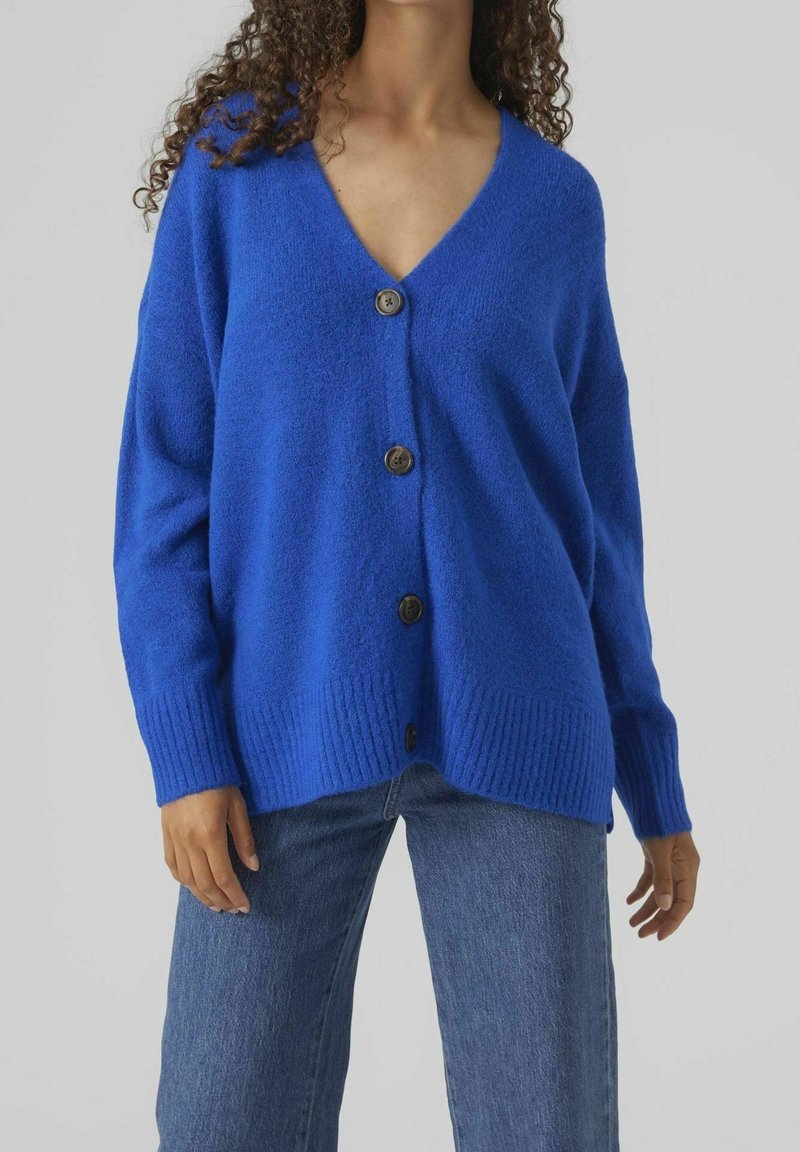 Femme portant un cardigan boutonné bleu vif et ample avec des poignets et un ourlet côtelés, associé à un jean bleu de lavage moyen.