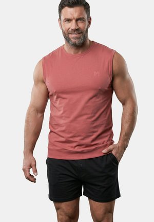 Muskelbepackter Mann lächelt, trägt ein ärmelloses rötliches Shirt und schwarze Shorts, steht mit einer Hand in der Hosentasche vor einem einfarbigen Hintergrund.