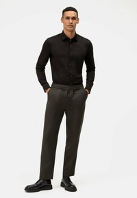 Zwarte langemouwenshirt met knopen, gecombineerd met donkergroene broek met elastische tailleband en zwarte veterschoenen.