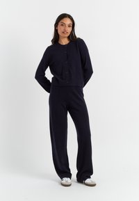 Marineblauwe gebreide cardigan met knopen aan de voorkant en bijpassende hoge taillebroek, met een ontspannen pasvorm en zachte textuur. Casual instap schoenen.