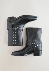 Botines de piel negra con tacón bajo, adornados con tachuelas plateadas y un detalle de correa con hebilla. Textura suave y diseño aerodinámico.
