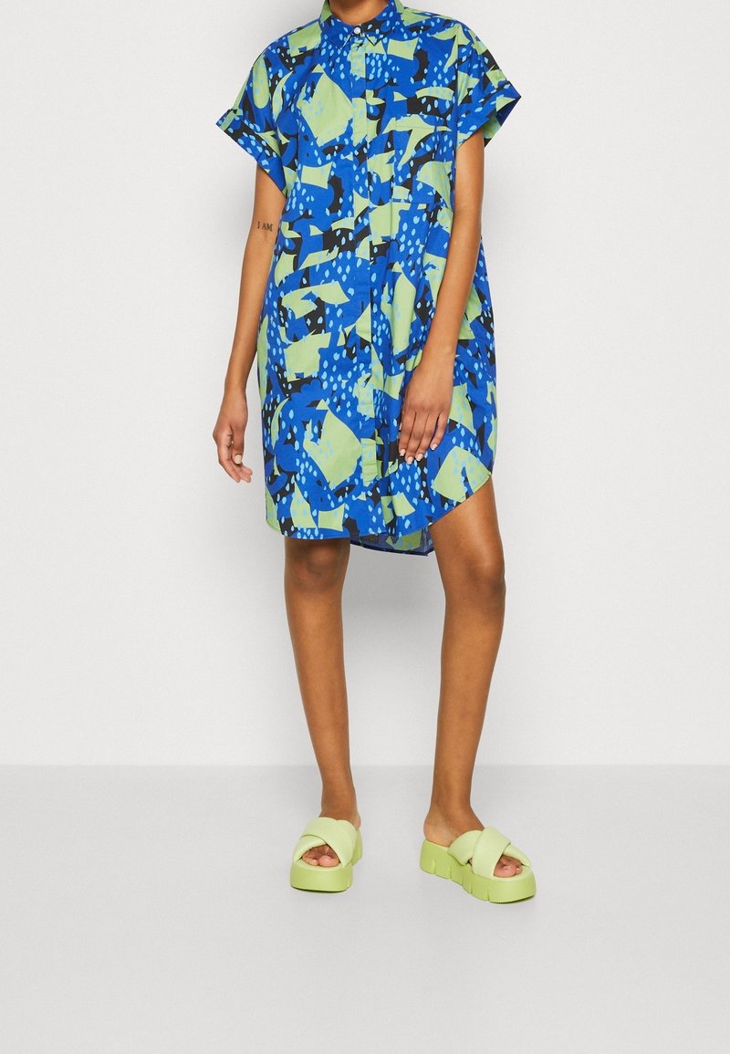 Robe à manches courtes bleue avec des motifs abstraits verts ; elle comporte un col, une fermeture à boutons sur le devant, et une coupe décontractée. Associée à des sandales à plateforme vert clair.