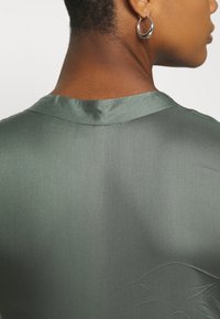 Satin grünes Oberteil mit glatter Textur, ausgestattet mit einem klassischen Rundhalsauschnitt und dezenten Designelementen auf der Rückseite.