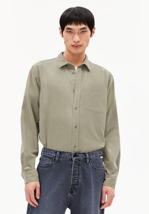 Chemise à manches longues vert olive clair avec un devant à boutons, une poche poitrine et une texture douce, associée à un jean en denim foncé.