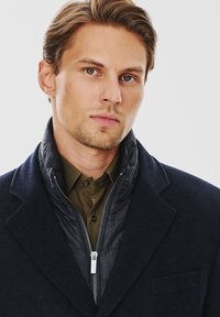 Granatowy wełniany płaszcz z wysokim kołnierzem, noszony na ciemnozielonej koszuli z guzikami. Posiada zapinaną na zamek podszewkę o pikowanej teksturze.