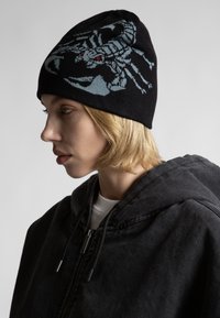 Căciulă tricotată neagră cu un grafic de scorpioni în detalii albastre deschise și roșii. Purtați-o cu o jachetă neagră cu glugă, care are un fermoar.
