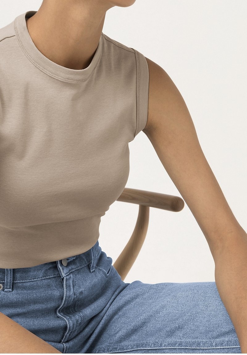 Light beige sleeveless top with a high neckline, made of soft cotton, paired with high-waisted blue denim jeans. Smooth texture, fitted design.