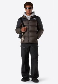 Puffer jacket in dark brown and black, featuring a zip front, insulated design, and a hood. Paired with black pants and sunglasses.
