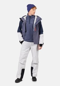 Man som bär en blå mössa, en marinblå och grå jacka, en marinblå fleece-tröja, vita snöbyxor med svarta lappar och svarta skor, står med händerna i fickorna.