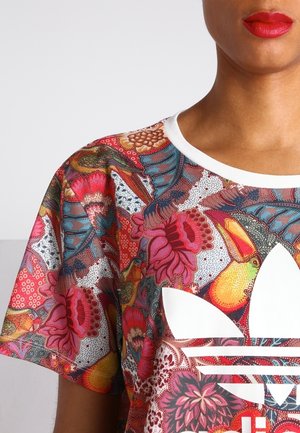 Persoon draagt een kleurrijk overhemd met bloemen- en toekanpatroon, met een groot wit Adidas-logo op de borst en rode lippenstift.