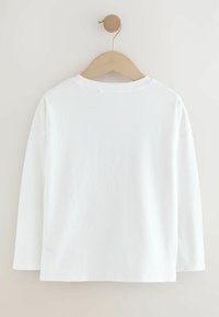 Una camiseta blanca de manga larga exhibida en un perchero de madera. Presenta un cuello redondo y un corte relajado, con una textura de tela suave y lisa.