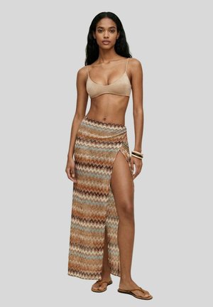 Donna che indossa un crop top beige e una gonna lunga con spacco alto, fantasia nei toni marrone, blu e beige, con braccialetti e sandali.