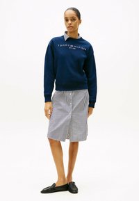 Felpa blu navy con logo bianco, abbinata a una gonna a righe con bottoni. Mocassini neri completano l'outfit. Texture dei tessuti lisci visibili.