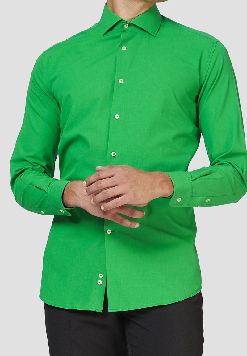 Homme portant une chemise boutonnée à manches longues vert vif avec des boutons beige, debout les mains jointes devant, sur un fond uni.